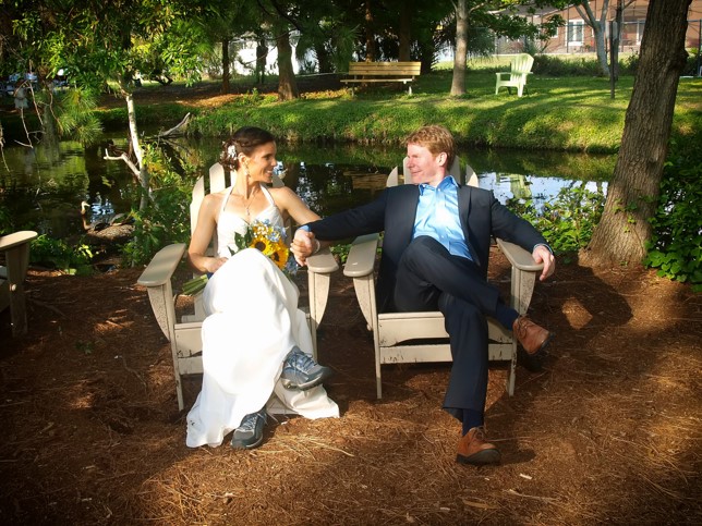 William Miller in a suit sits outdoors holding hands with Kim Clairy in a wedding dress and bouquet, near a pond.