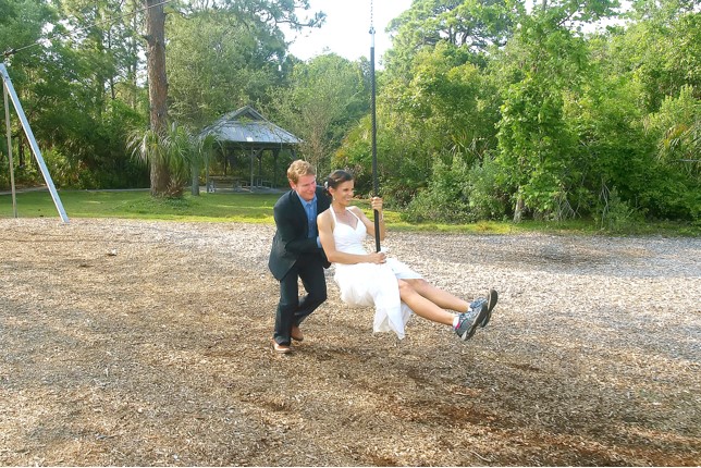 Kim Clairy in a wedding dress on a rope swing, being pushed by her husband in a suit in an outdoor setting.