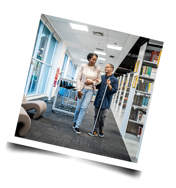 An adult assisting another adult who is using a white cane in a library