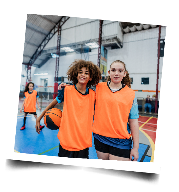 2 teens in orange practice jerseys stand smiling together in a gym, 1 holding a basketball, 1 with their arm around the other's shoulder