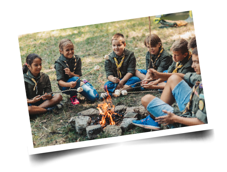 A group of kids in scout uniforms sitting around a campfire toasting marshmallows on sticks